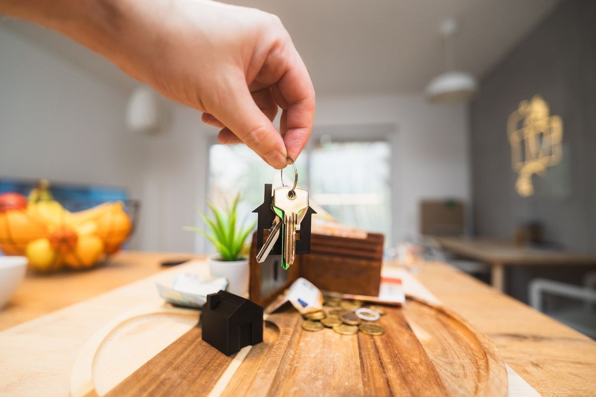 Hand holding house keys above a tray with a small house model, wallet, and coins