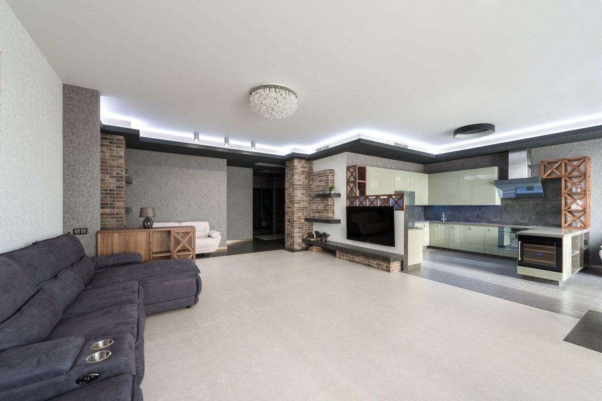 Open-plan modern apartment with a grey sectional sofa, brick feature wall with TV, and kitchen beyond